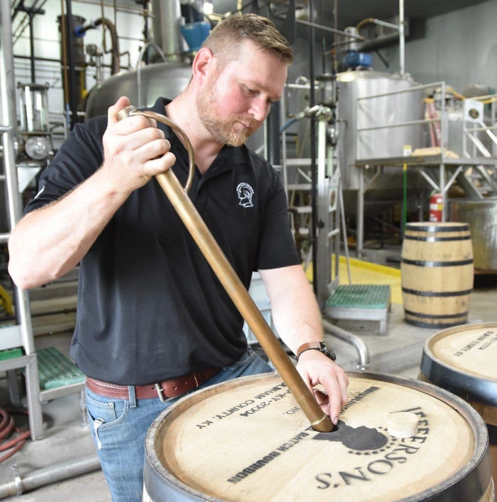 A distiller drawing a sample from a barrel with a whiskey thief